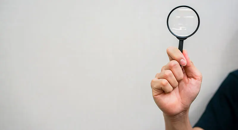 Hand holding a small magnifying glass against a plain grey background.