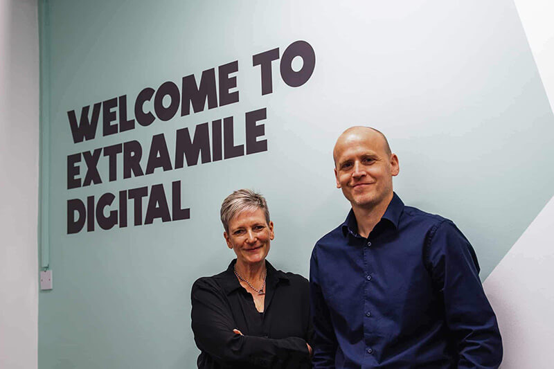 Welcome to ExtraMile Digital sign with two smartly dressed colleagues below